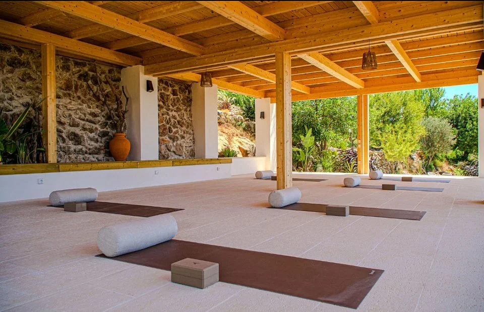 Open-air covered studio with mats, bolsters, and blocks set out beside a stone wall and garden