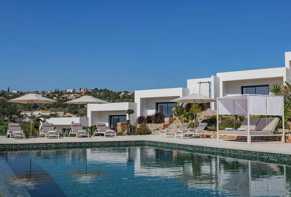 Pool with sun loungers, umbrellas, and a shaded daybed in front of white villas
