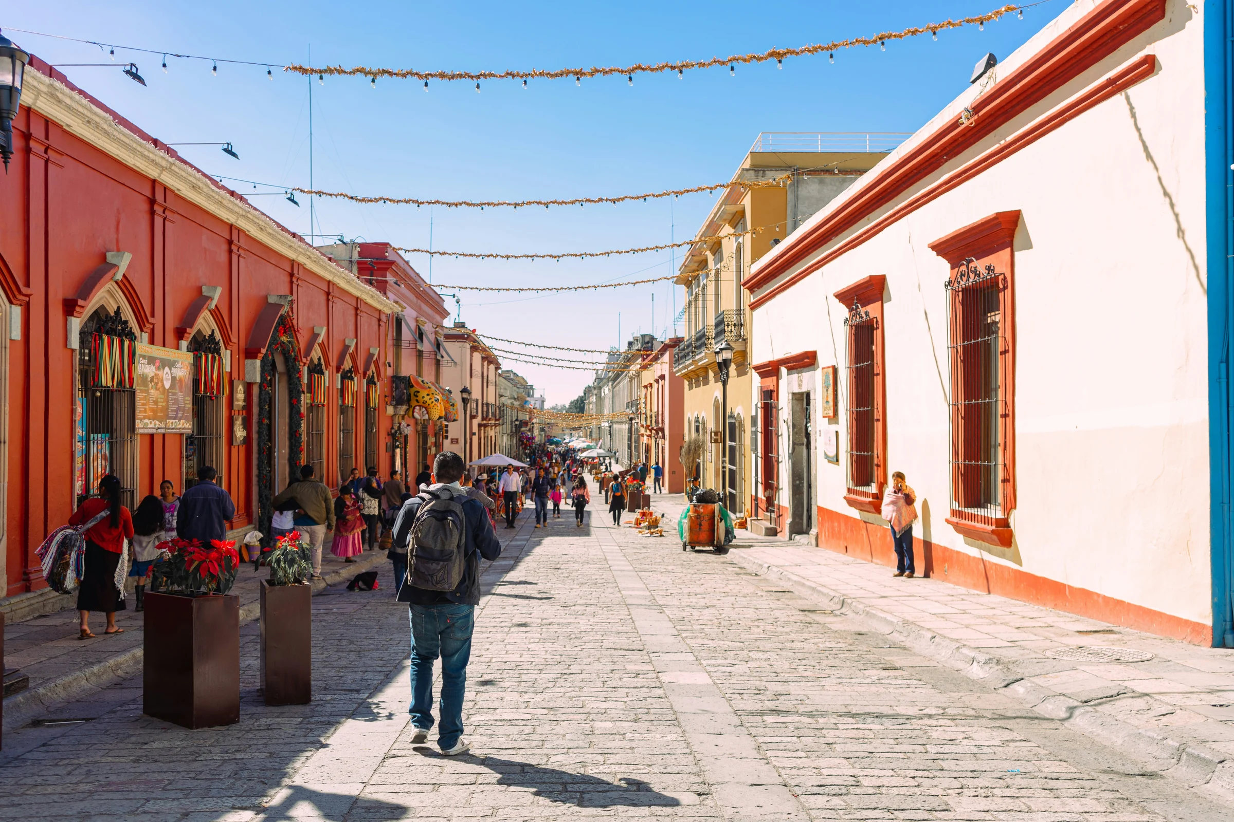 People walking down a sunny pedestrian street in Oaxaca's historic center, lined with orange and white colonial buildings