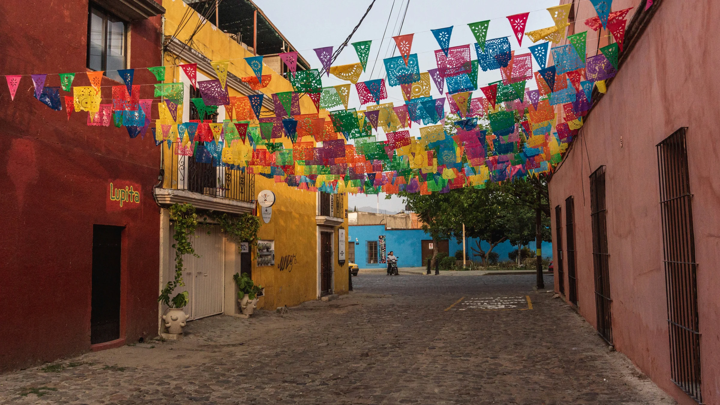 Colorful papel picado banners strung over a cobblestone street lined with red, yellow and pink buildings in Oaxaca